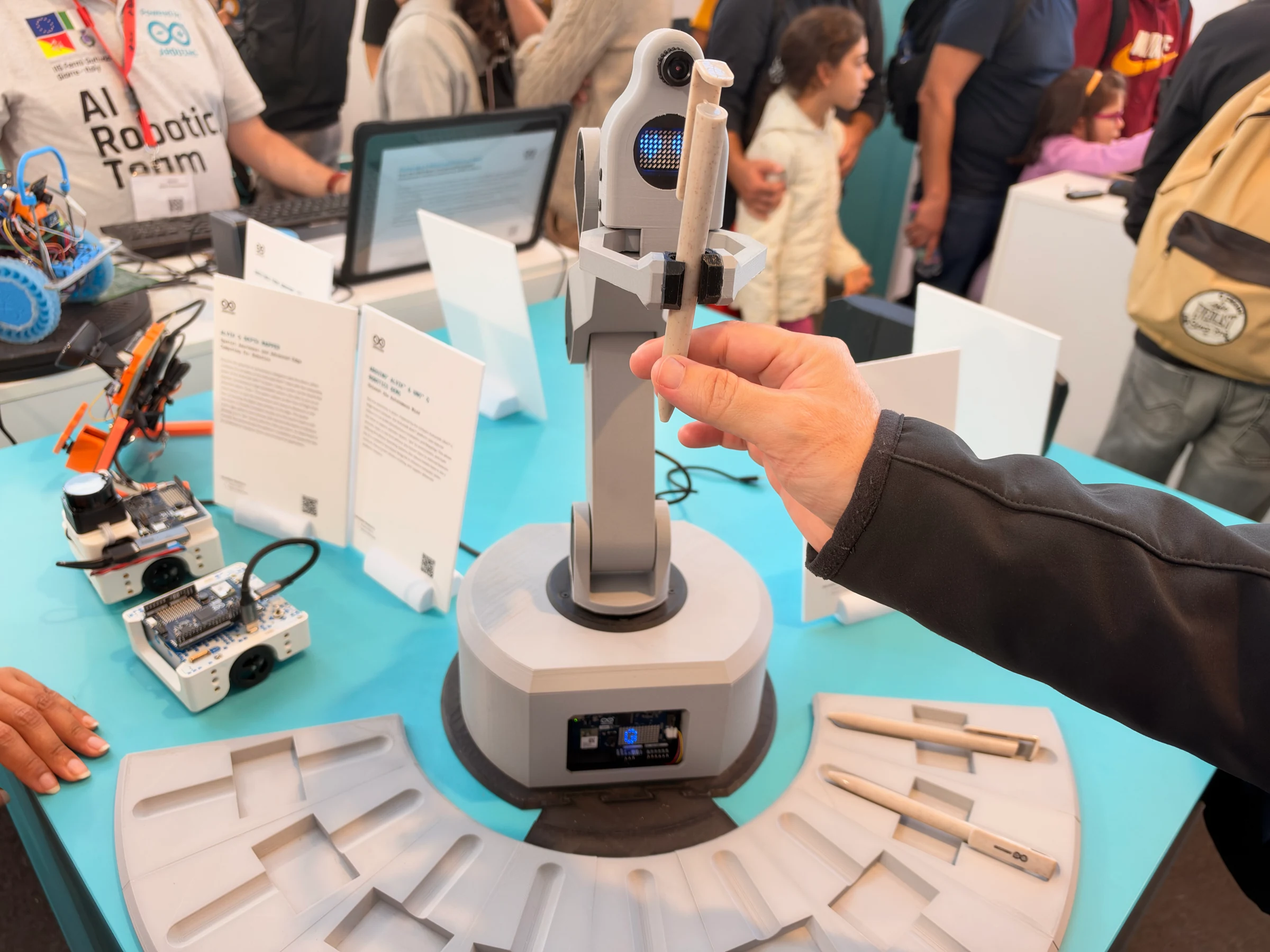 A visitor takes a pen held by a gray desktop robot arm with a camera, LED-matrix face, and circular pen organizer.