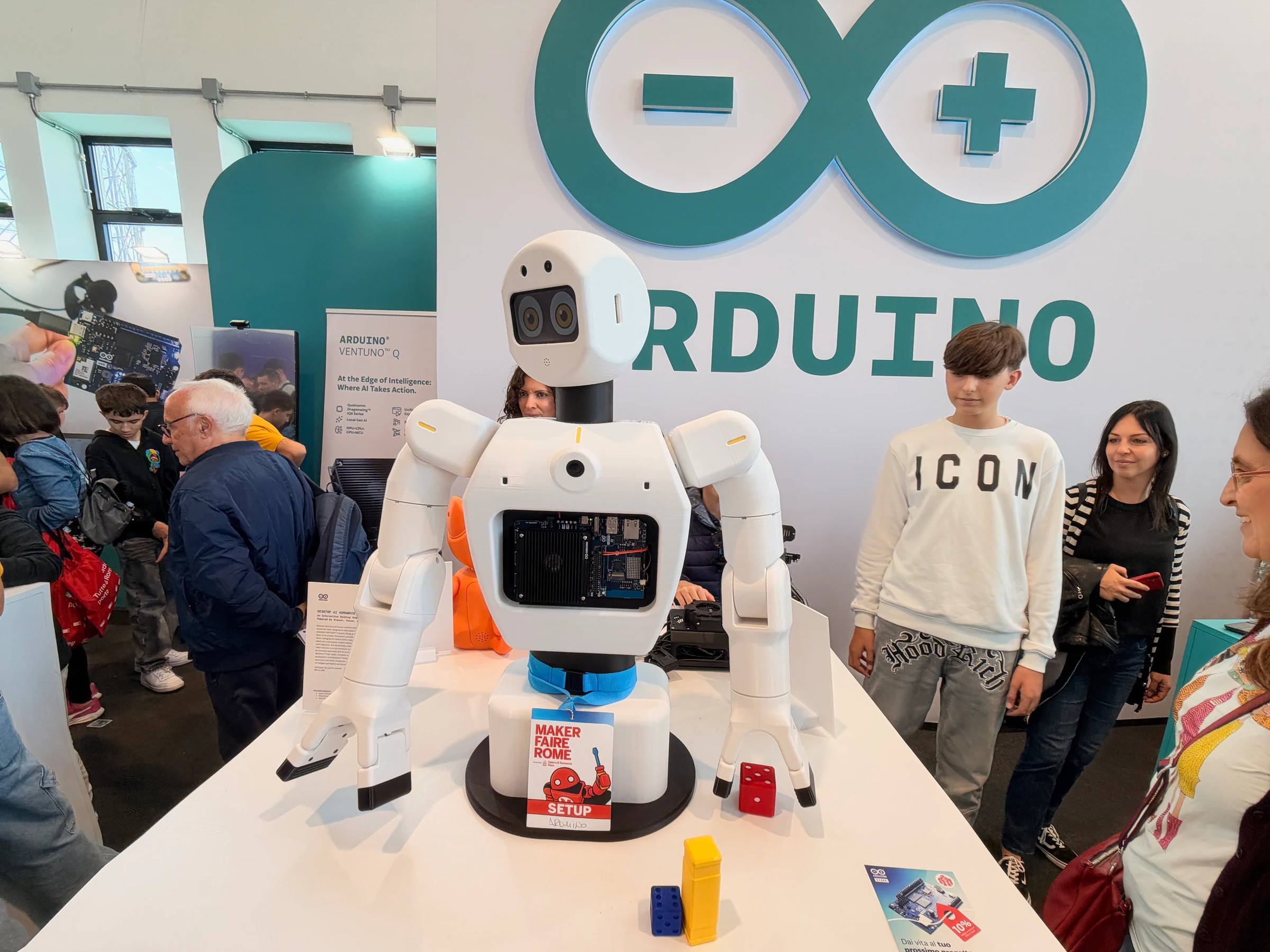 White desktop humanoid robot at the Arduino booth with animated eyes, articulated arms, and an exposed computing board in its chest.