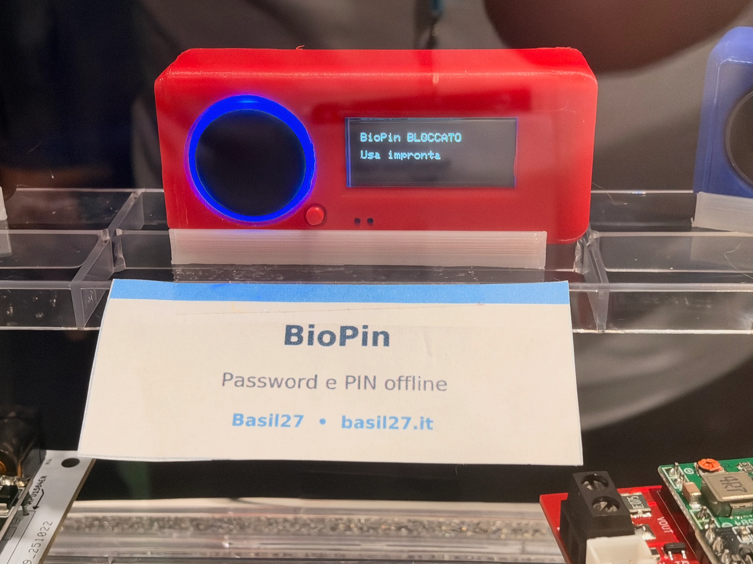 Red Basil27 BioPin device with a blue-lit fingerprint sensor and a screen asking the user to unlock it with a fingerprint.