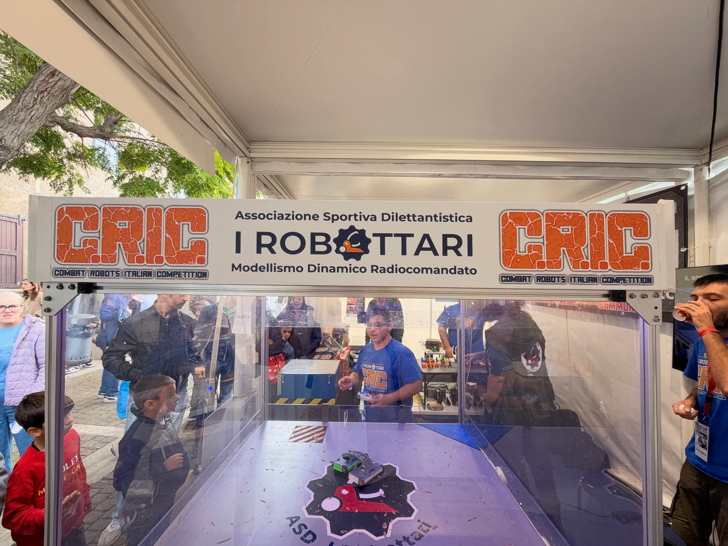 Visitors watch small combat robots inside the transparent I Robottari arena beneath a Combat Robots Italian Competition banner.