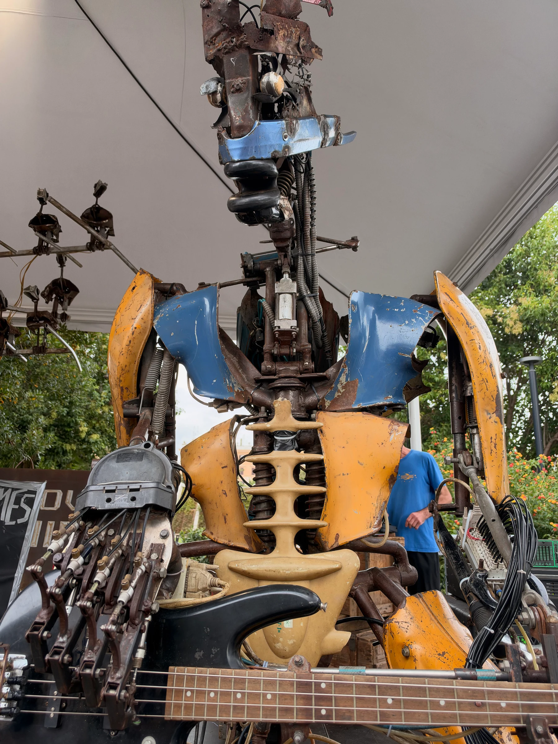 Scrap-metal robot bass player with blue and yellow body panels, exposed mechanisms, and metal fingers positioned over a bass guitar.