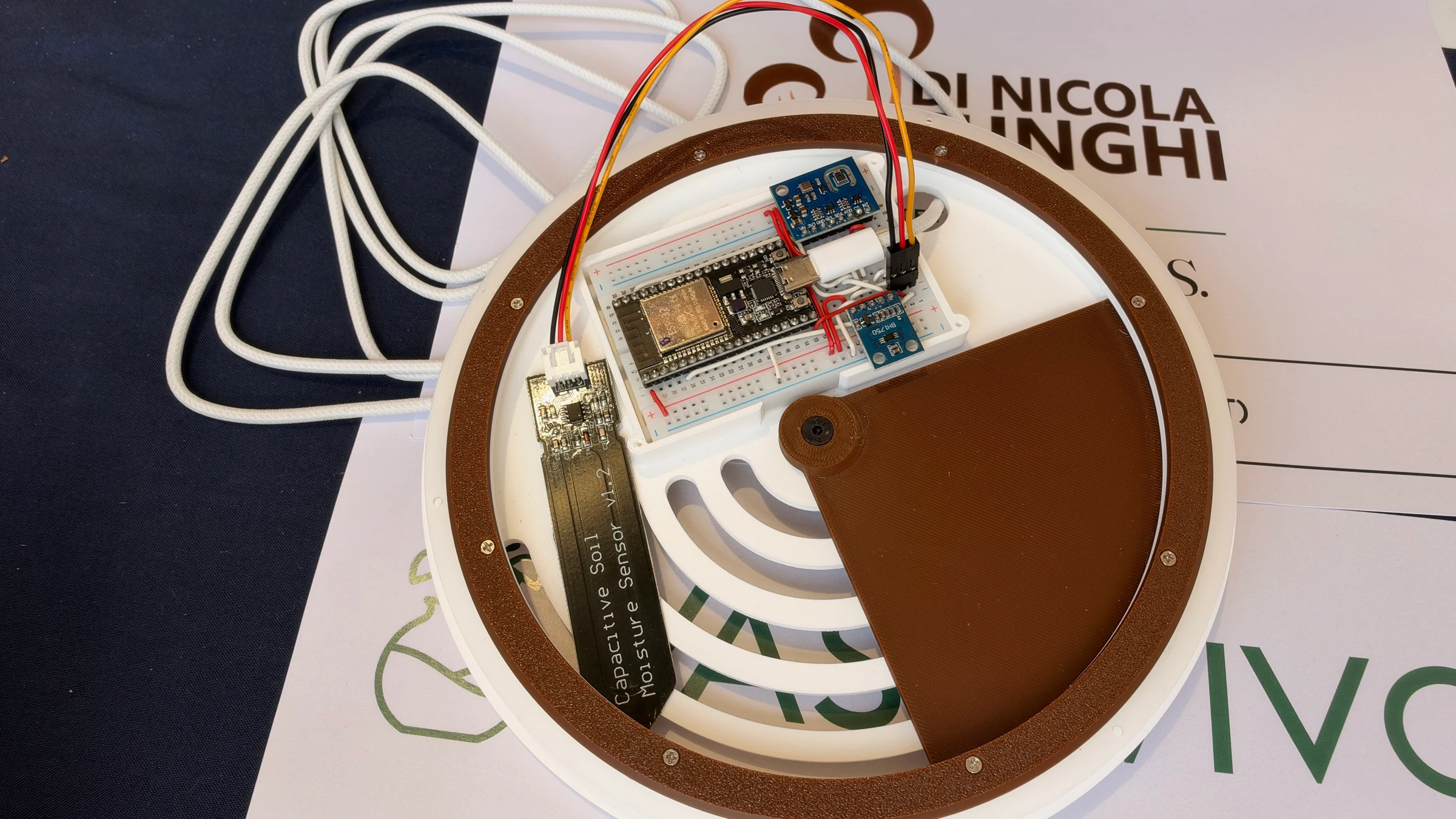 Underside of a VasoVivo lid showing an ESP32 board, breadboard, blue sensor modules, wiring, and a capacitive moisture probe.
